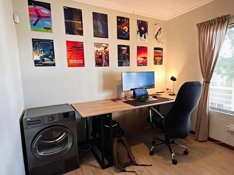 A functional home office space featuring laminate flooring, a window with blinds, and a built-in laundry appliance integrated into the room layout.