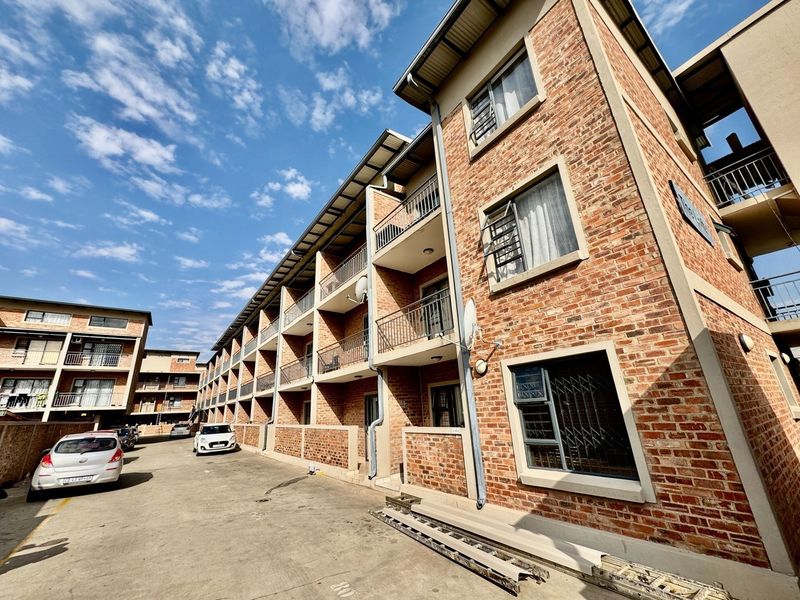 Exterior view of a multi-story apartment complex featuring face brick facade, private balconies with metal railings, and a paved driveway area.
