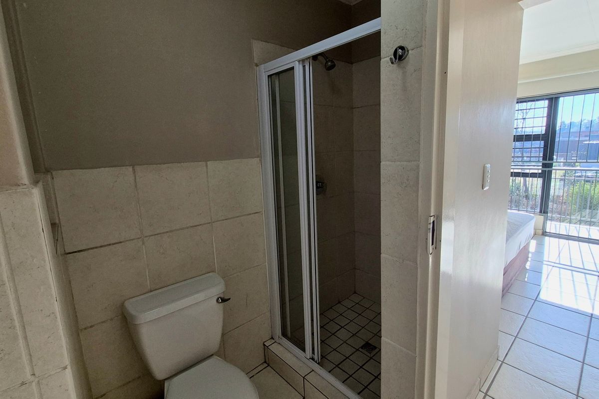 A functional bathroom featuring a shower enclosure and toilet, with neutral wall and floor tiling, adjacent to a bright living area.