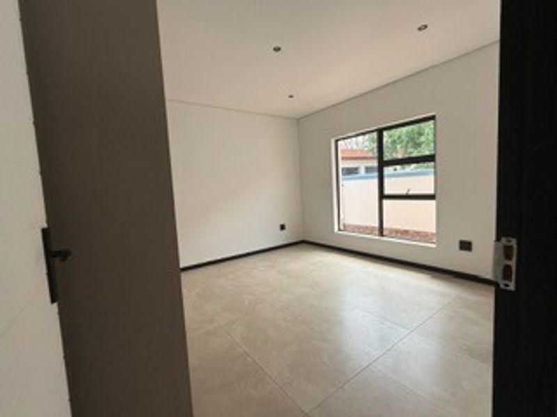 A bright, empty bedroom featuring neutral tiled flooring, recessed downlighting, and a large aluminium-framed window.