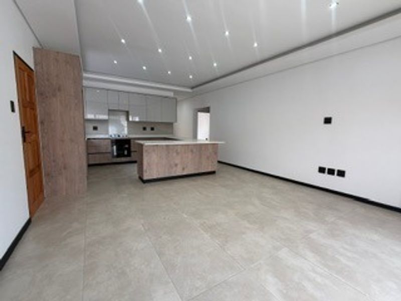 A modern, open-plan living and kitchen area featuring neutral tiled flooring, contemporary cabinetry, a central kitchen island, and recessed ceiling lighting.