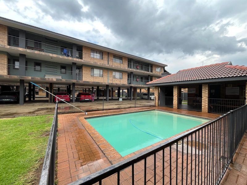 Communal swimming pool area featuring brick paving, safety fencing, and a covered pavilion, situated within an apartment complex with undercover parking.