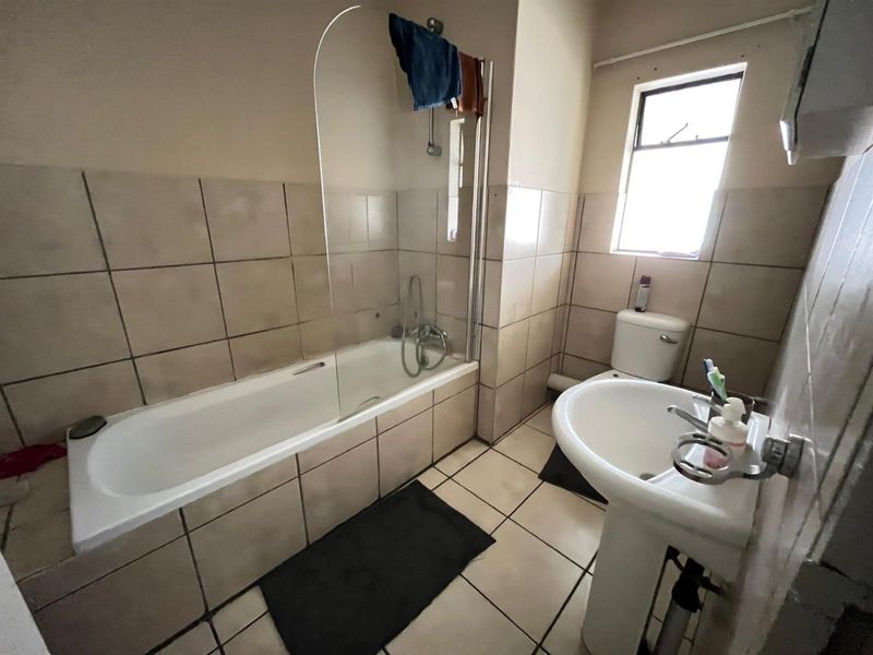 A dated bathroom featuring a bathtub with a glass screen, pedestal sink, and toilet, finished with neutral ceramic wall and floor tiles.