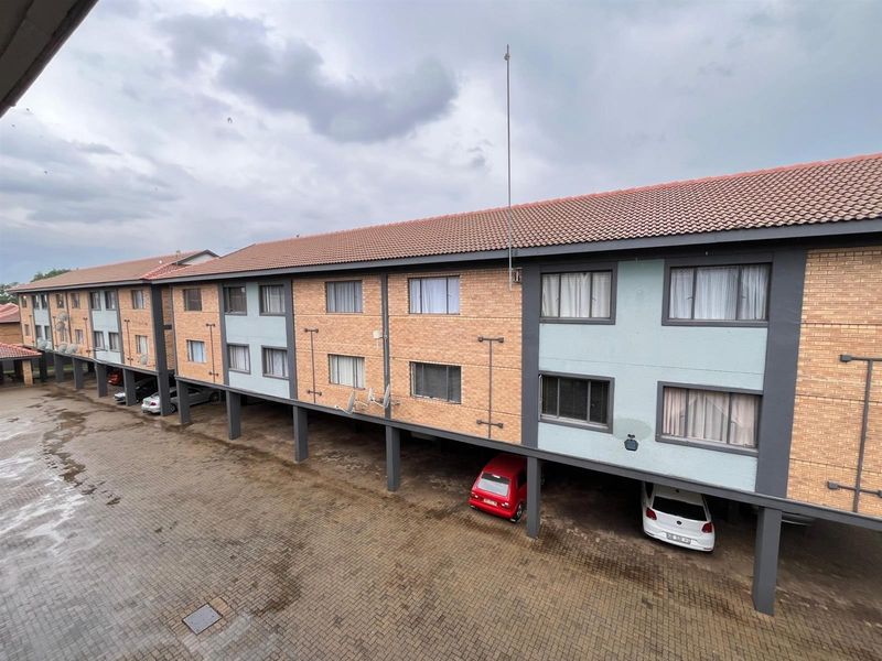 Exterior view of a multi-story apartment complex featuring brick and painted facade, tiled roof, and ground-level covered parking bays with paved driveway.