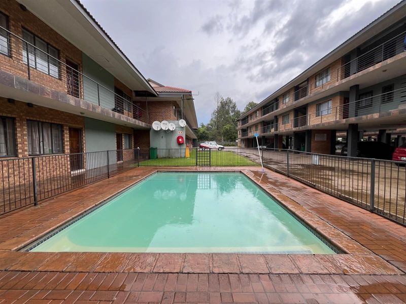 Communal swimming pool area situated between two multi-story apartment blocks, featuring brick paving and secure metal perimeter fencing.