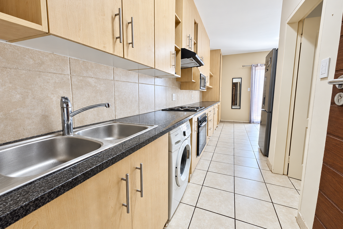 A functional galley-style kitchen featuring light wood cabinetry, granite countertops, integrated appliances, and neutral ceramic floor tiling.