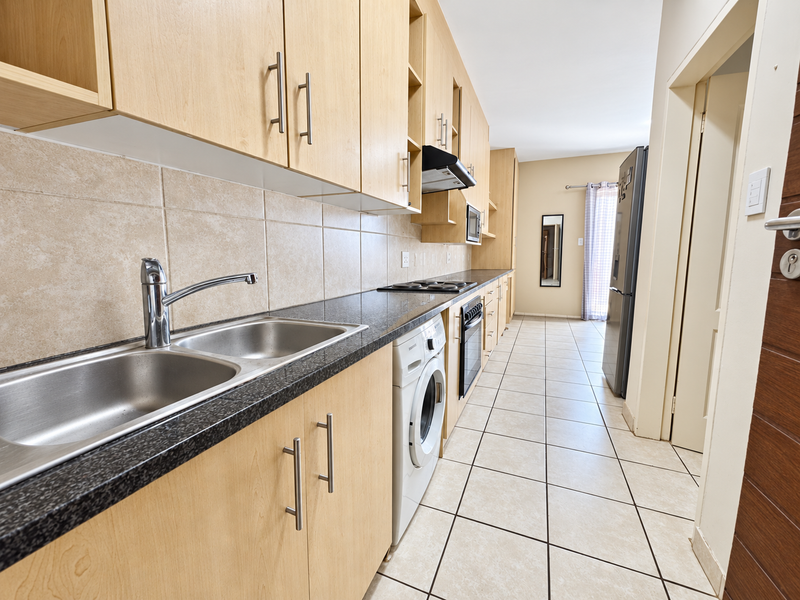 A functional galley-style kitchen featuring light wood cabinetry, granite countertops, integrated appliances, and neutral ceramic floor tiling.