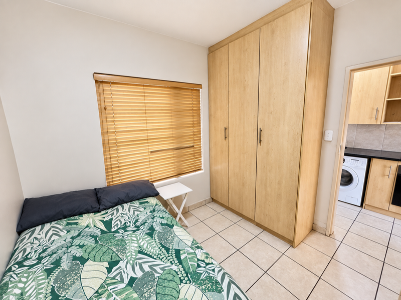 A well-maintained bedroom featuring light wood-finish built-in wardrobes, tiled flooring, and venetian blinds, with an adjacent view into the kitchen area.