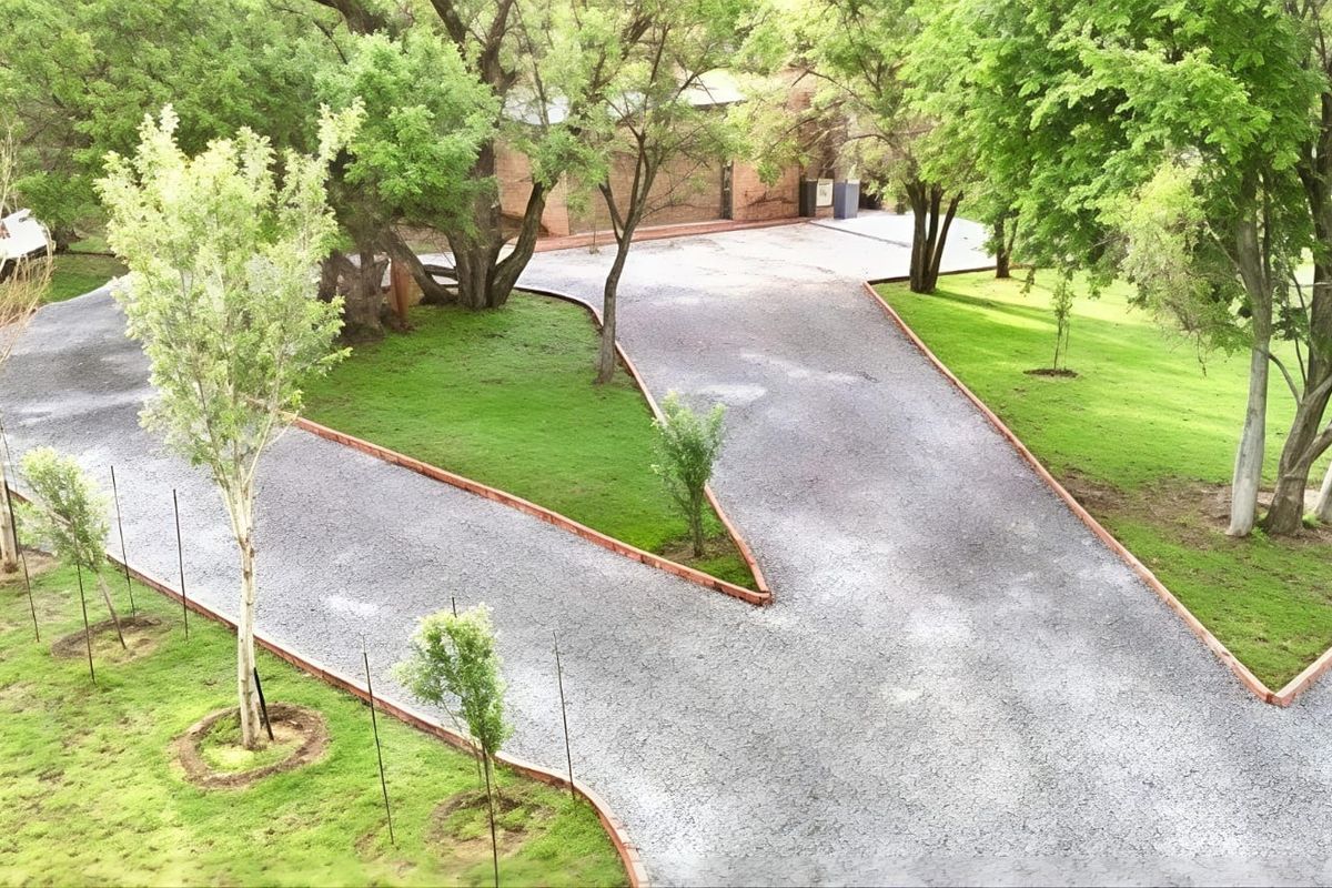 An aerial view of a property featuring a well-maintained gravel driveway with brick edging, surrounded by lush green lawns and mature trees.