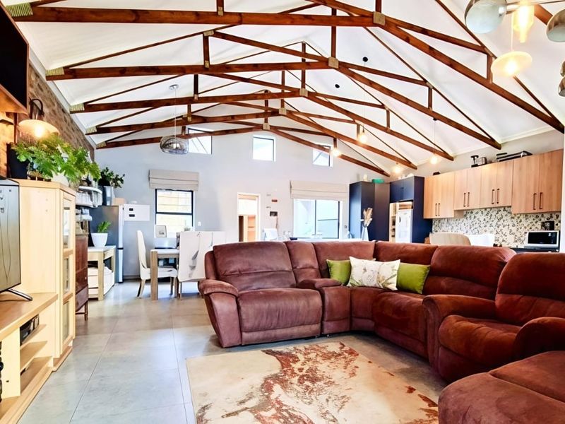 Spacious open-plan living and kitchen area featuring high vaulted ceilings with exposed timber trusses, tiled flooring, and a feature exposed brick wall.