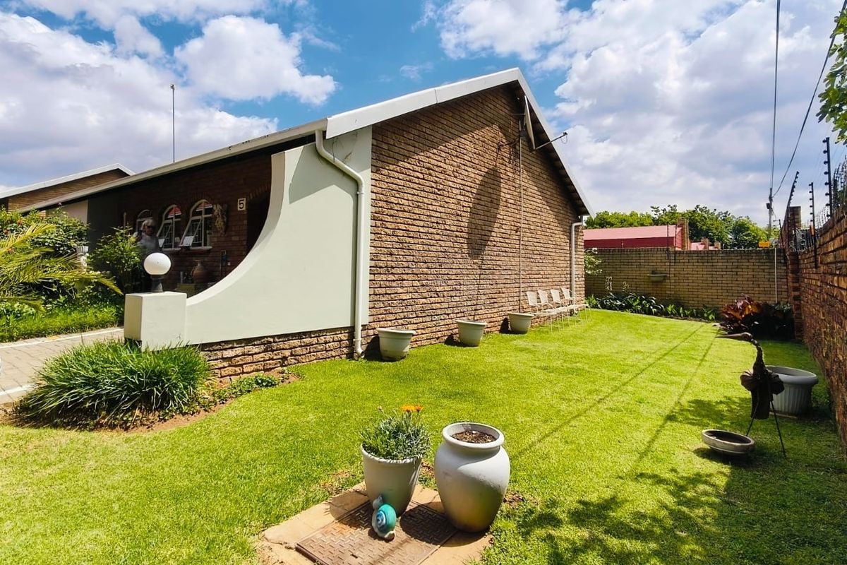 A well-maintained rear exterior view of a face brick townhouse featuring a manicured lawn, paved walkway, and secure perimeter wall with electric fencing.