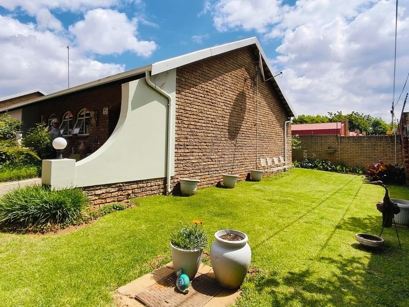 A well-maintained rear exterior view of a face brick townhouse featuring a manicured lawn, paved walkway, and secure perimeter wall with electric fencing.