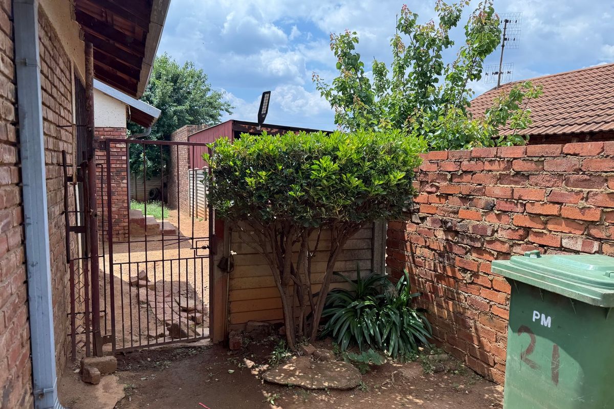 A rear exterior view showing a brick boundary wall, metal security gate, and basic garden area with established shrubbery.