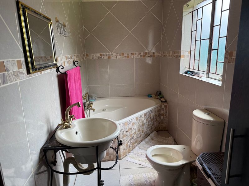 A compact bathroom featuring a corner bathtub, pedestal basin, and toilet with tiled walls and security bars on the window.