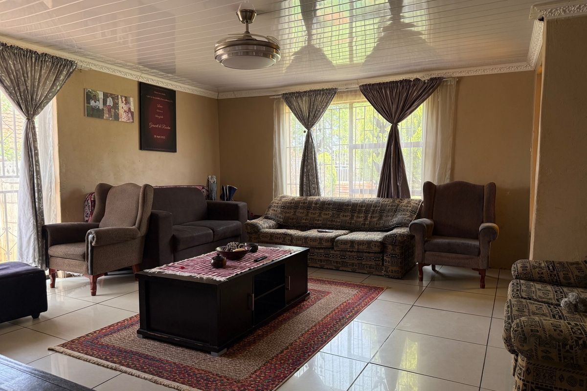 A spacious living room featuring tiled flooring, a ceiling fan, and large windows with security bars, offering a functional and comfortable living space.
