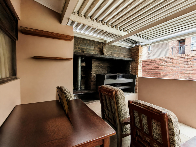 Covered patio area featuring a built-in brick braai, corrugated metal roofing, and floating wall shelves, providing a functional outdoor entertainment space.