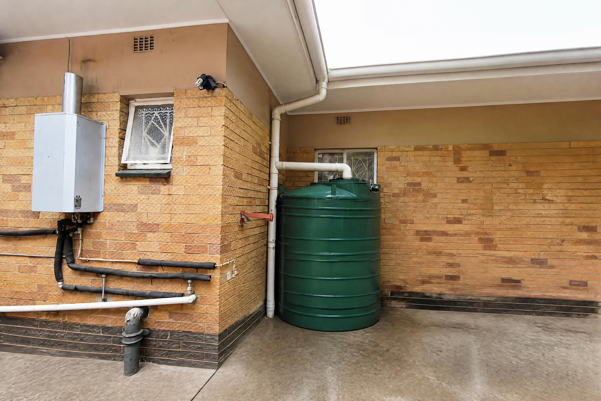 Rear exterior area featuring a brick facade, gas geyser installation, and a large green water storage tank connected to the roof drainage system.