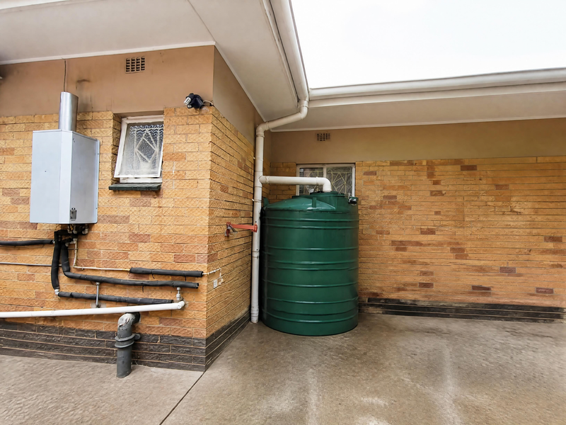 Rear exterior area featuring a brick facade, gas geyser installation, and a large green water storage tank connected to the roof drainage system.