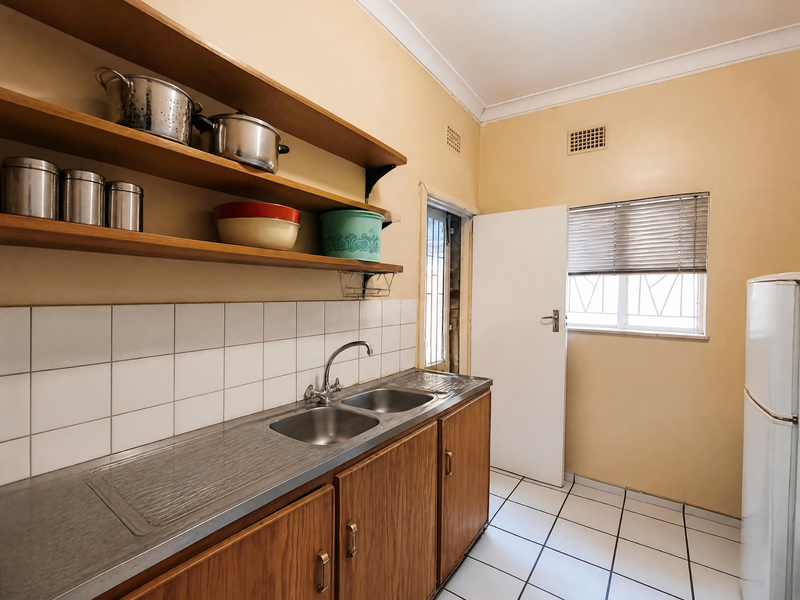 A functional kitchen featuring wooden cabinetry, open shelving, a double stainless steel sink, and tiled flooring with a neutral wall finish.