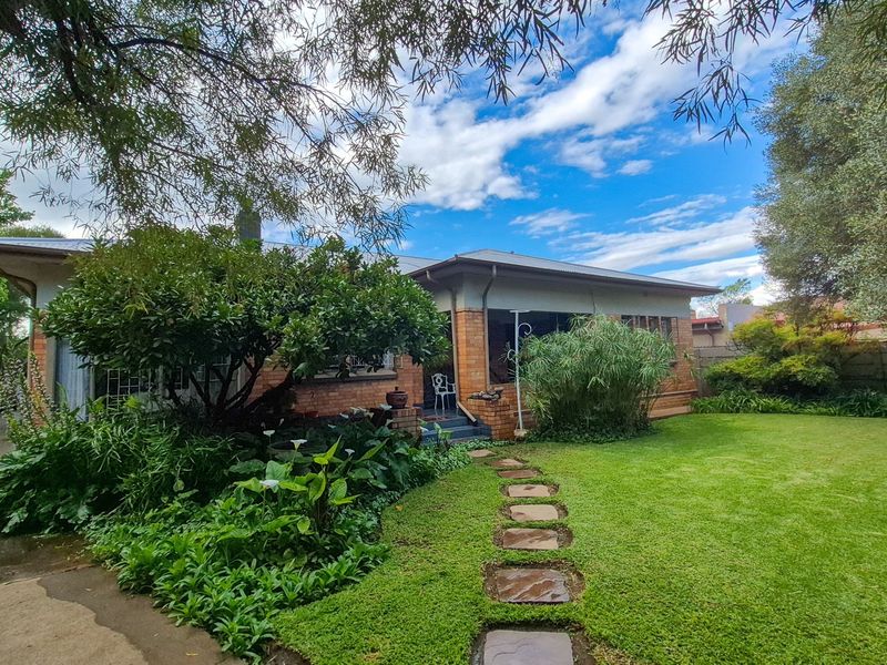 A charming rear view of a brick house featuring a covered patio, a well-maintained lawn, and a stone pathway leading through a lush garden.