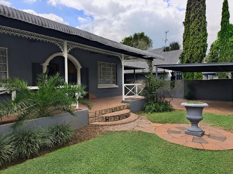 Charming front exterior featuring a covered veranda with decorative ironwork, stone steps, a well-maintained garden, and a paved driveway with a carport.