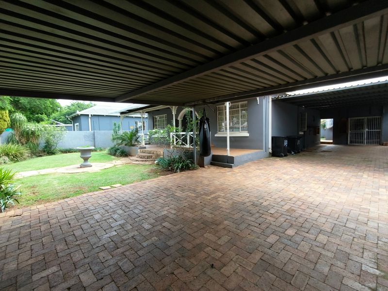 Spacious rear exterior featuring a large paved driveway, covered carport, and a covered patio area overlooking a landscaped garden.