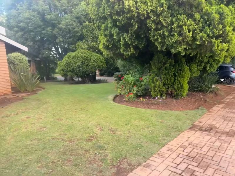 A well-maintained front garden featuring a paved walkway, established trees, and manicured lawn areas adjacent to the brick house exterior.