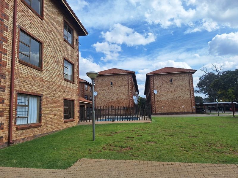 Exterior view of a multi-story face brick apartment complex featuring landscaped lawn areas, paved walkways, and secure perimeter fencing.