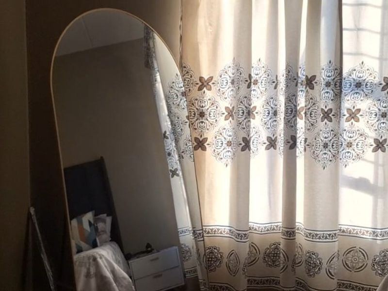 A bedroom interior featuring a window with patterned curtains and a large arched floor mirror.