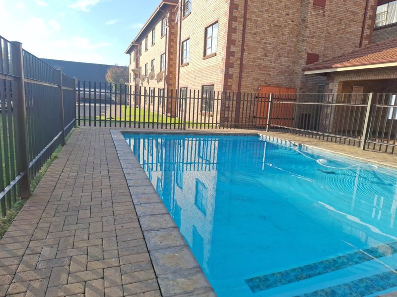 A communal swimming pool area featuring a paved deck and secure metal perimeter fencing, set against a brick apartment building.