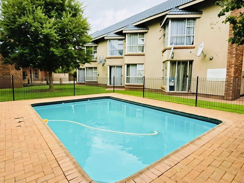 A well-maintained communal swimming pool area featuring brick paving, secure metal fencing, and views of the apartment building exterior.
