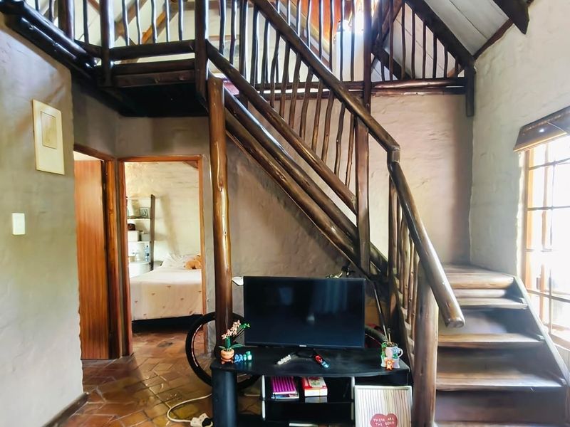 Living area featuring a rustic wooden staircase, exposed timber rafters, and terracotta floor tiles, leading to an adjacent bedroom.