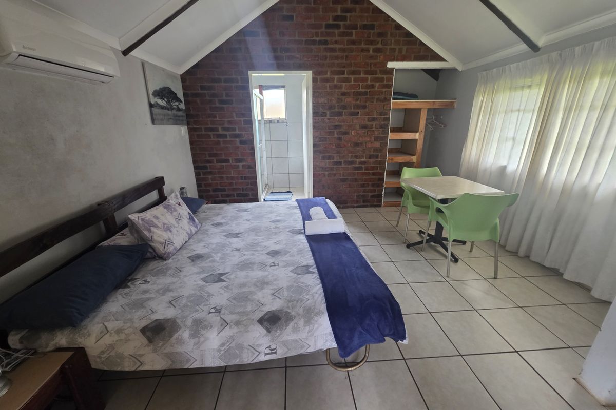 A bedroom featuring a vaulted ceiling, exposed brick feature wall, tiled flooring, and an en-suite bathroom entrance.