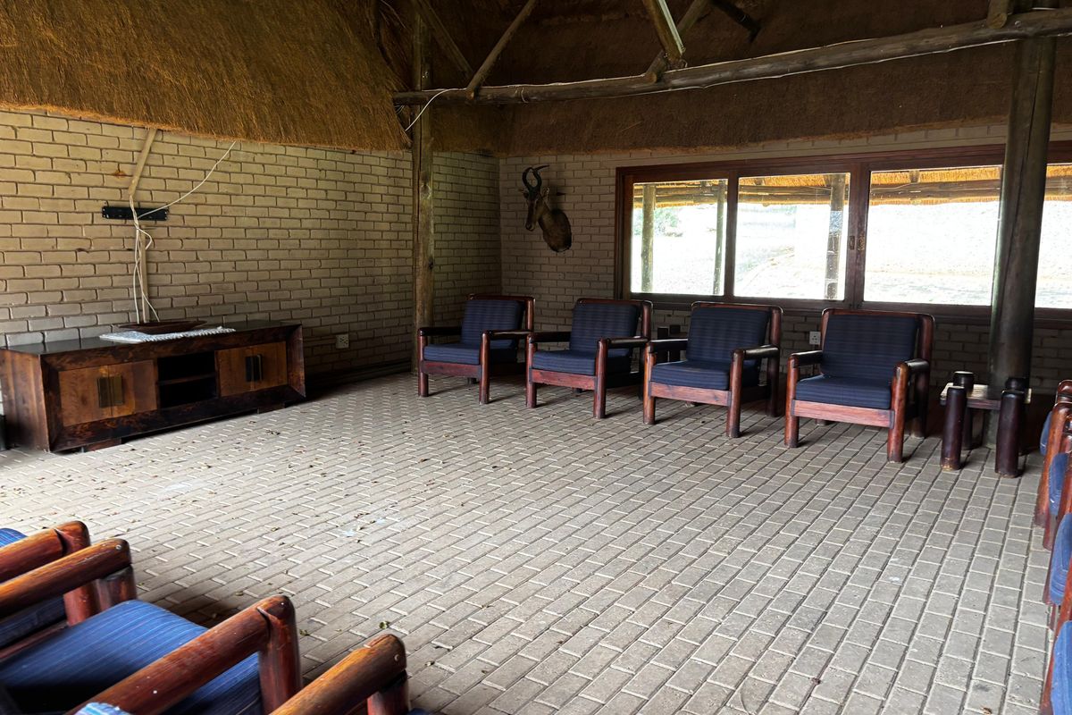 Spacious lodge living area featuring a high thatched roof, exposed timber beams, brick walls, and paved flooring with large windows.