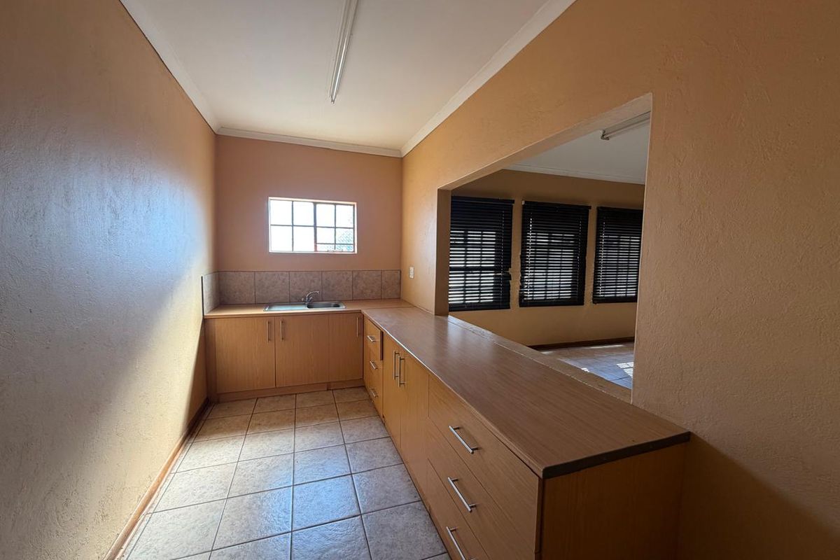 A functional kitchen area featuring built-in wooden cabinetry, laminate countertops, a stainless steel sink, and tiled flooring, with an open-plan serving hatch to the living area.
