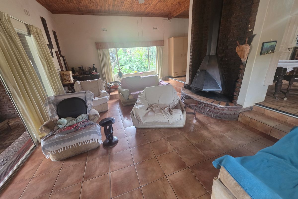 Spacious living room featuring a prominent brick fireplace, exposed timber ceiling, tiled flooring, and large windows providing natural light and garden views.