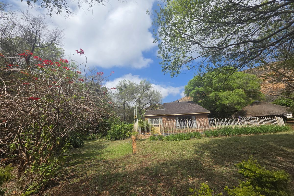 A view of the rear brick farmhouse elevation featuring a tiled roof, metal fencing, and an adjacent thatched lapa set within a garden.