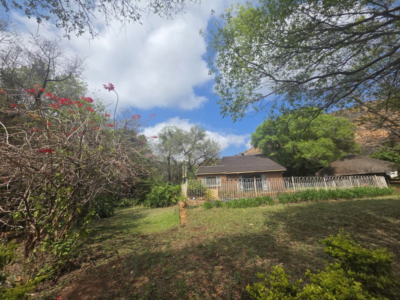 A view of the rear brick farmhouse elevation featuring a tiled roof, metal fencing, and an adjacent thatched lapa set within a garden.