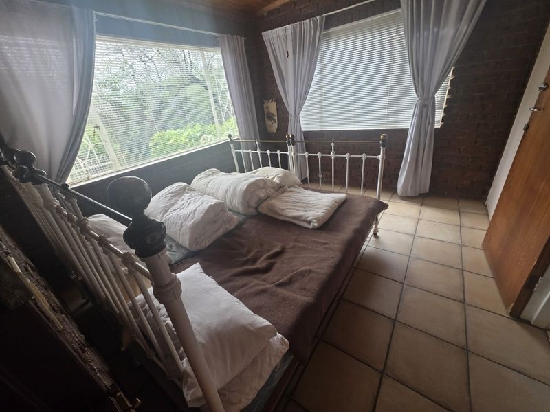 A rustic bedroom featuring exposed brick walls, tiled flooring, and large windows with venetian blinds, offering a cozy and character-filled living space.