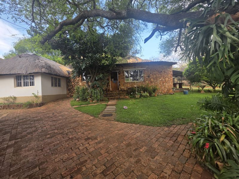 A charming farm-style residence featuring traditional stone cladding, a thatched roof section, and a brick-paved driveway set within a lush, mature garden.