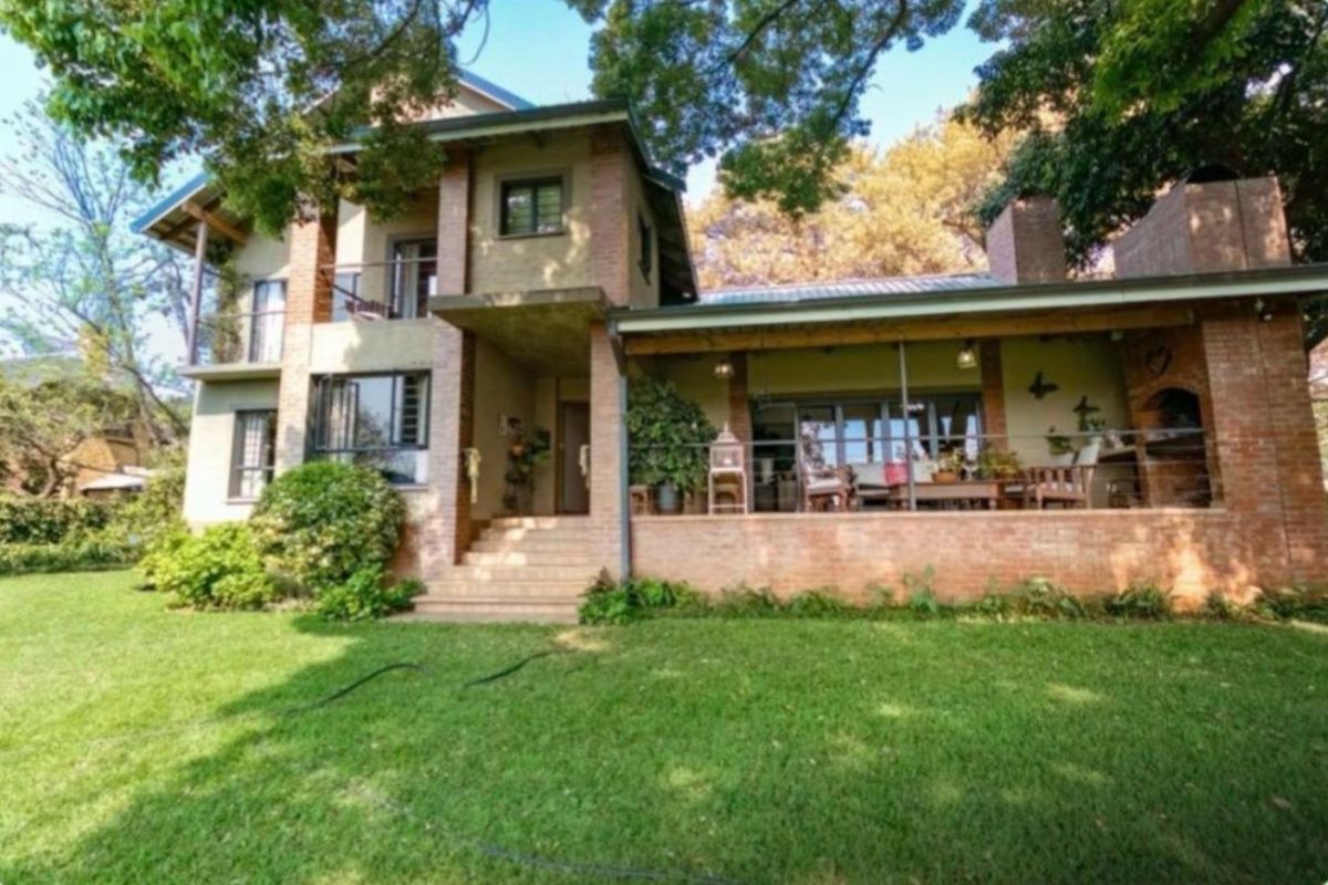 A multi-story brick residence featuring a spacious covered patio with a built-in braai, overlooking a well-maintained garden and mature trees.