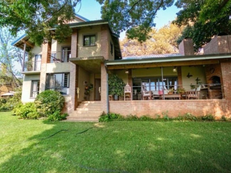A multi-story brick residence featuring a spacious covered patio with a built-in braai, overlooking a well-maintained garden and mature trees.
