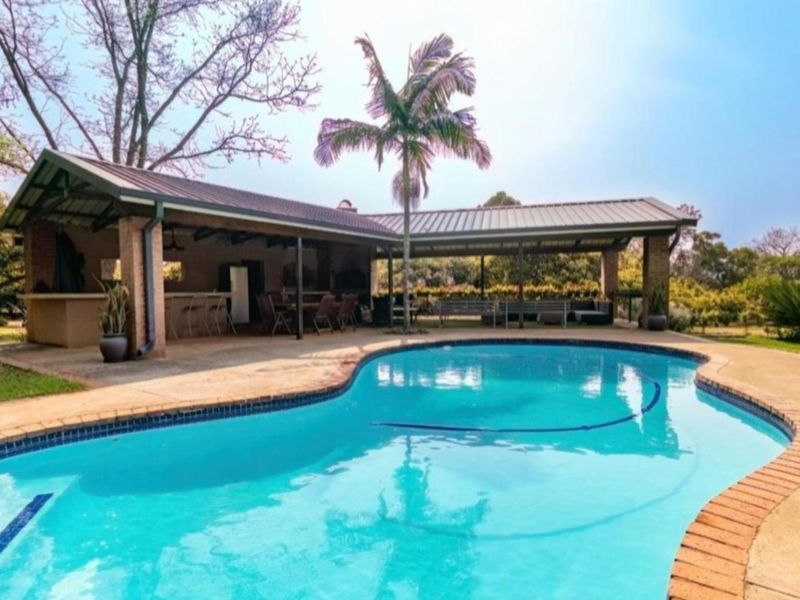 Spacious outdoor entertainment area featuring a large swimming pool, covered patio with built-in braai, and established garden surroundings.