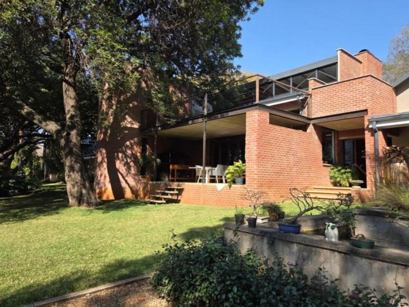Rear view of a multi-level face-brick residence featuring a covered patio, an upper-level balcony, and a landscaped garden with a concrete retaining wall.