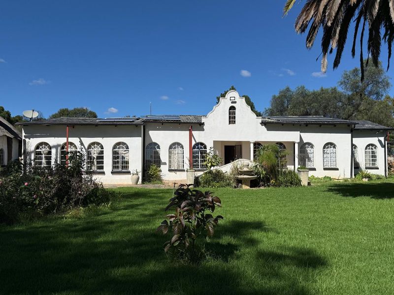 A charming farm-style residence featuring a white facade with arched windows, a gabled roof, solar panels, and a central stone fountain in the garden.