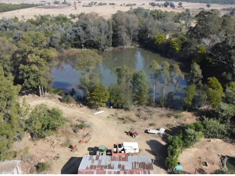 Aerial view of a farm property featuring a large dam, surrounding woodland, and outbuildings with corrugated metal roofing.