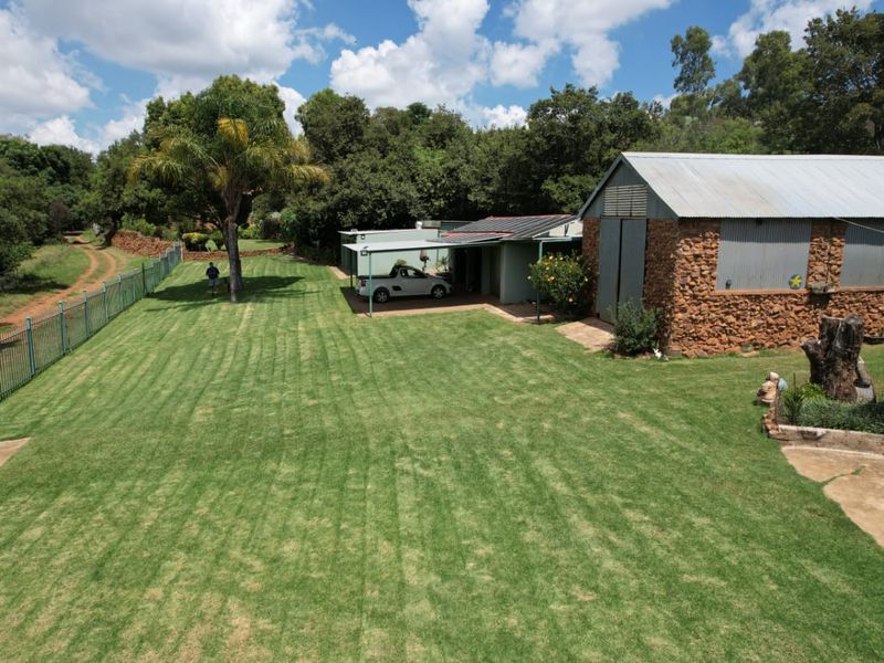 An elevated view of a spacious backyard featuring a well-maintained lawn, a stone-clad outbuilding with a metal roof, and a covered carport area.