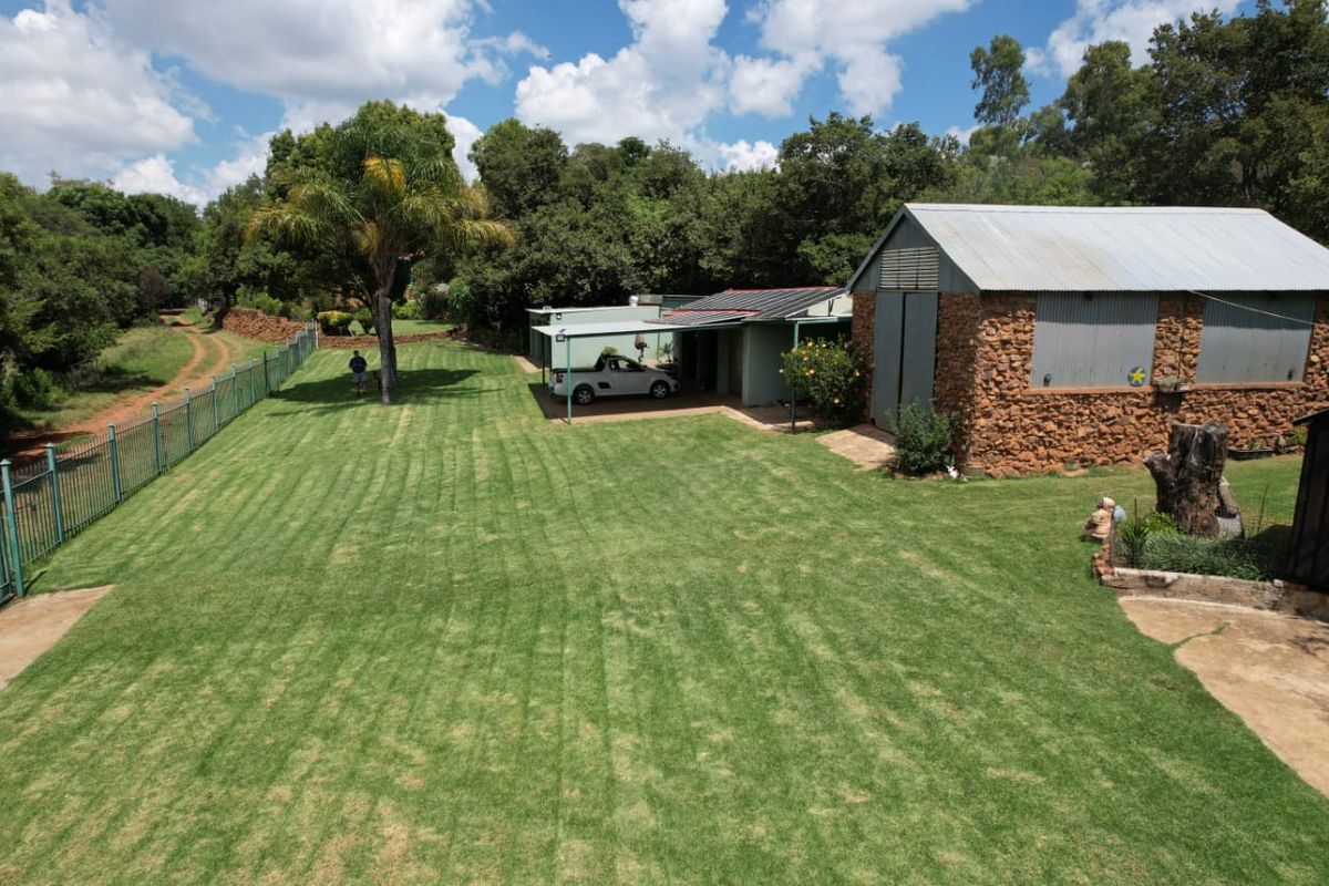 An elevated view of a spacious backyard featuring a well-maintained lawn, a stone-clad outbuilding with a metal roof, and a covered carport area.
