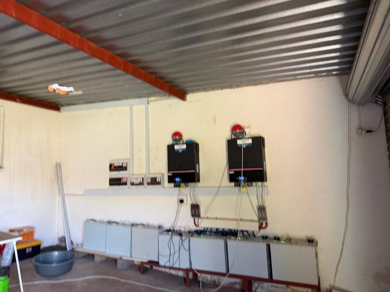 Utility space featuring a solar power installation with dual inverters and a battery storage bank mounted against a corrugated metal ceiling structure.