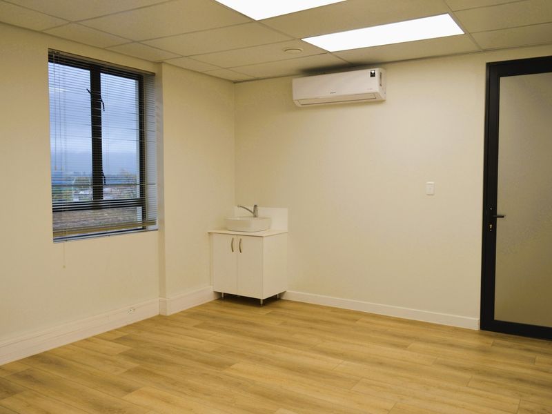 A clean, modern office space featuring laminate flooring, a built-in vanity with basin, split-system air conditioning, and large windows with blinds.